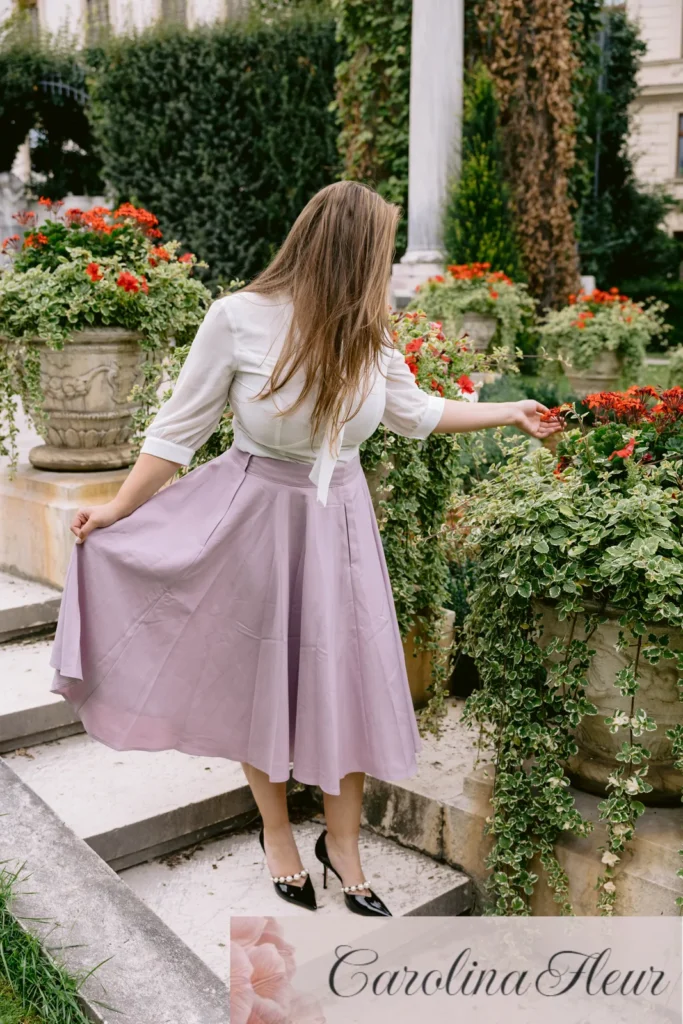 Carolina Fleur in weisser Bluse und fliederfarbenem Rock zwischen Blumen auf einer Steintreppe