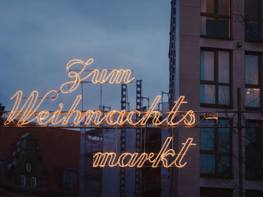 Leuchtschrift „Zum Weihnachtsmarkt“ in warmen Lichterketten vor einer Häuserfassade in der Abenddämmerung