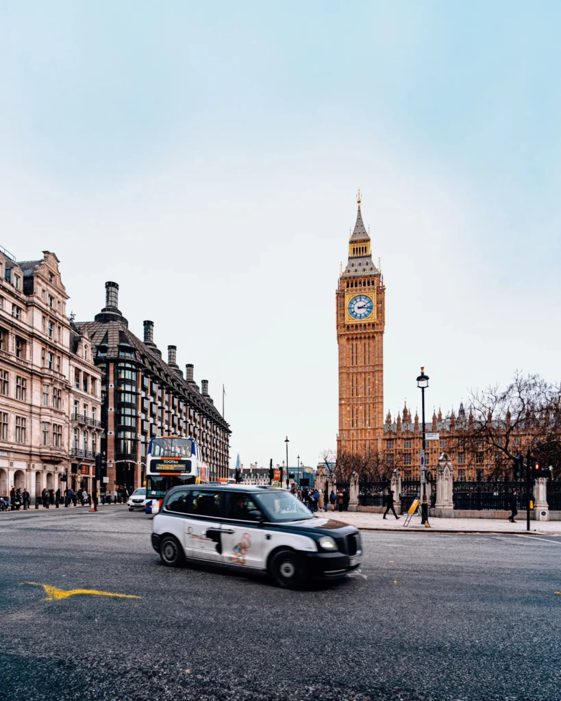 Big Ben und das Parlamentsgebäude in London, davor ein vorbeifahrendes Black Cab und ein Doppeldeckerbus